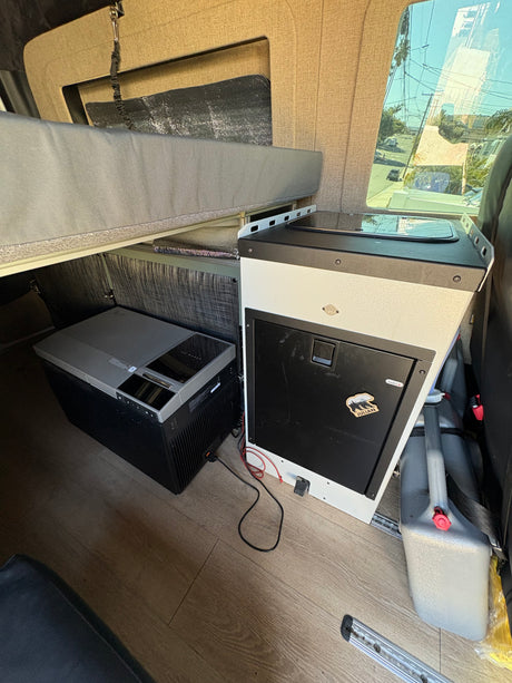 Inside an rv rental: compact camper van kitchen, portable fridge/ice maker, and raised bed setup for mobile living.
