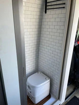Inside an RV rental: a compact, tiled wet bath featuring a composting toilet on a wooden mat and a towel rack.