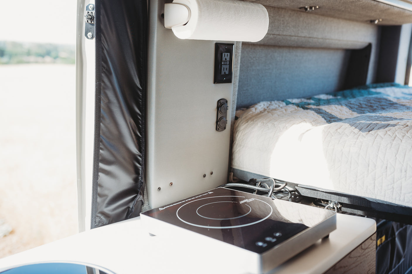 Inside an RV rental: a neatly organized space with a paper towel dispenser, outlets, induction cooktop, and a cozy bed.