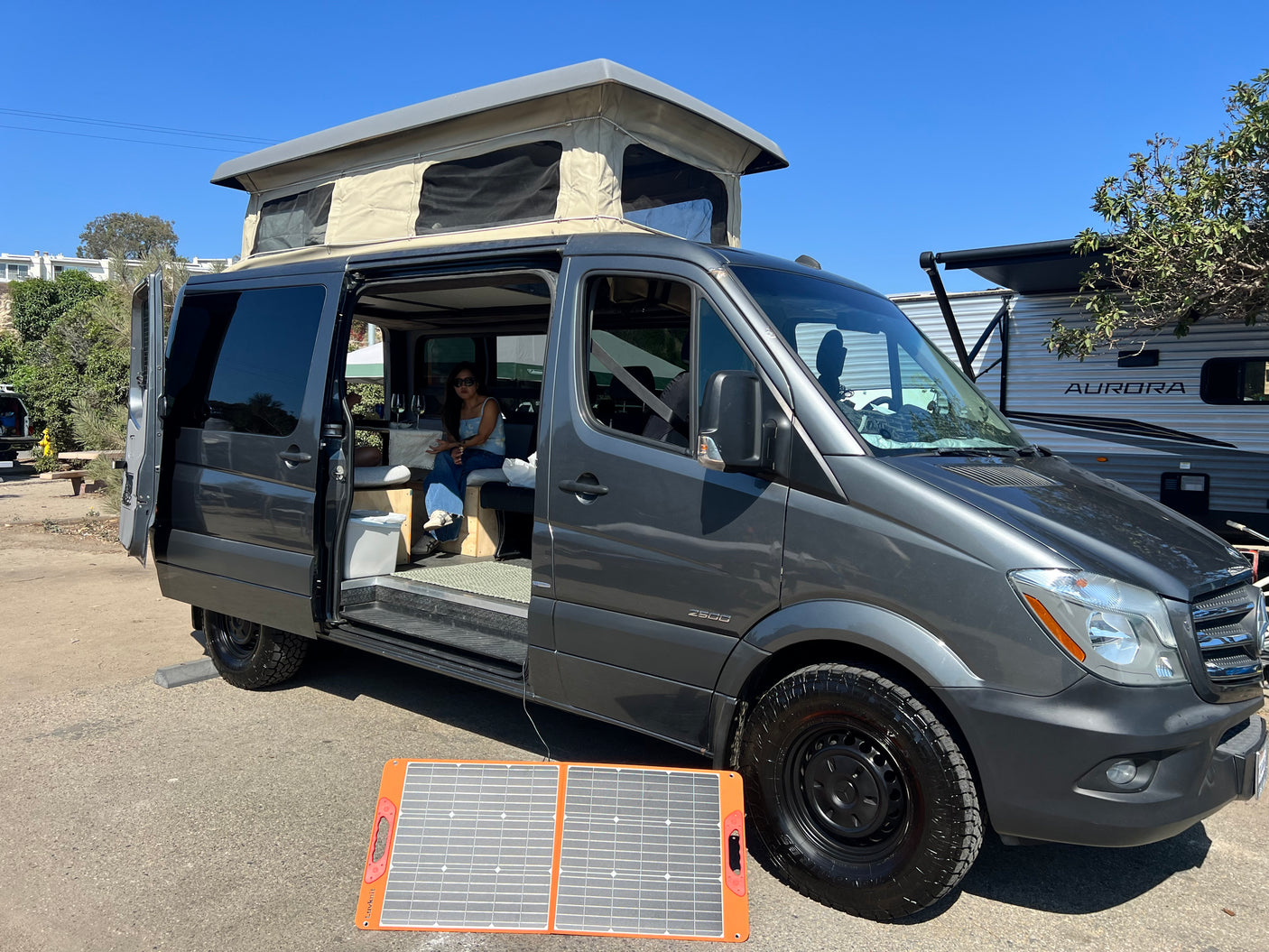 A grey pop-top Sprinter camper van, perfect for rv rental, uses solar power. A person relaxes inside the open door.