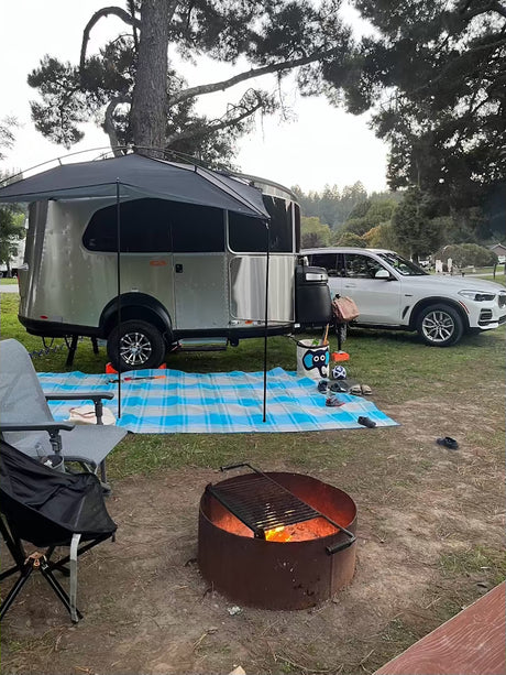 Cozy rv rental setup: a silver trailer with an awning & burning fire pit at a wooded campsite, ready for relaxation.