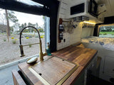 RV rental interior: A bright camper van kitchen with gold faucet, wood counter, & park view. Cozy bed in back.