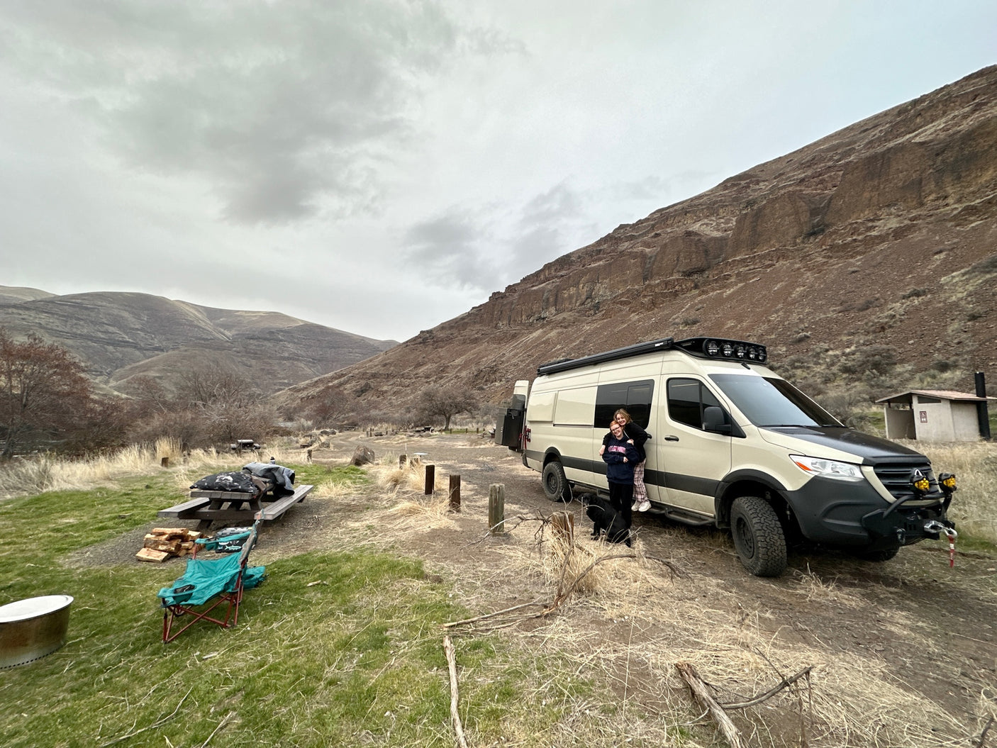 An rv rental parked at a scenic mountain campsite with people, a dog, and picnic setup under cloudy skies.