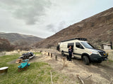 An rv rental parked at a scenic mountain campsite with people, a dog, and picnic setup under cloudy skies.
