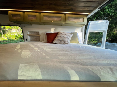 Bright camper van interior: large bed with pillows, olive overhead storage, wood ceiling, & open views for an rv rental.