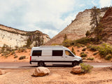 Scenic RV rental: A white Papago camper van parked on a dirt pullout with stunning layered desert mountains.