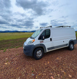 White Ram ProMaster camper van (RV rental) on a gravel road, backed by snowy mountains under cloudy skies. Explore the outdoors!