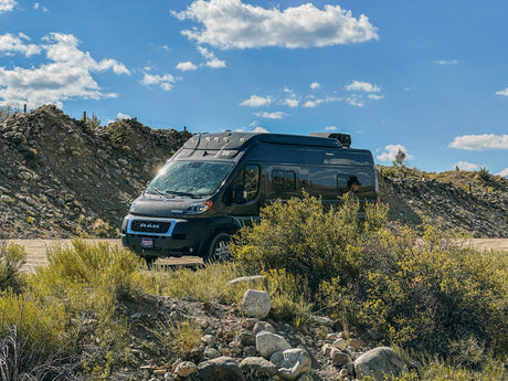 Adventure awaits with this RV rental! Dark camper van parked in a sunny, rocky outdoor setting, person in window.