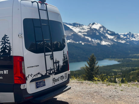 Back of a white Sprinter van RV rental with mountain decals, parked overlooking a scenic Montana lake and snowy peaks.