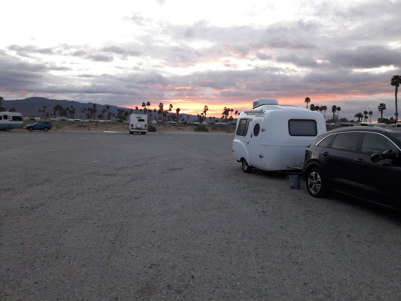 White compact RV rental and SUV parked on a gravel lot. Sunset glows over distant mountains and palm trees.