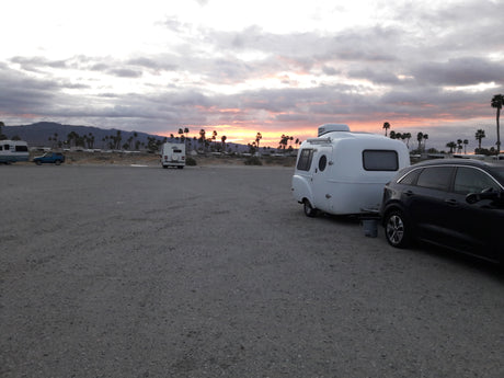 White compact RV rental and SUV parked on a gravel lot. Sunset glows over distant mountains and palm trees.
