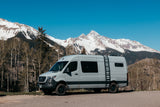 Scenic RV rental parked on a mountain road, with a grey camper van set against majestic snow-capped peaks.