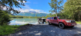 Red truck with an RV rental trailer backed by a serene lake and snow-capped mountains. Ideal for off-grid camping.