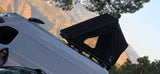 RV rental van with rooftop tent & smiling person, ready for adventure amidst scenic mountains and trees.