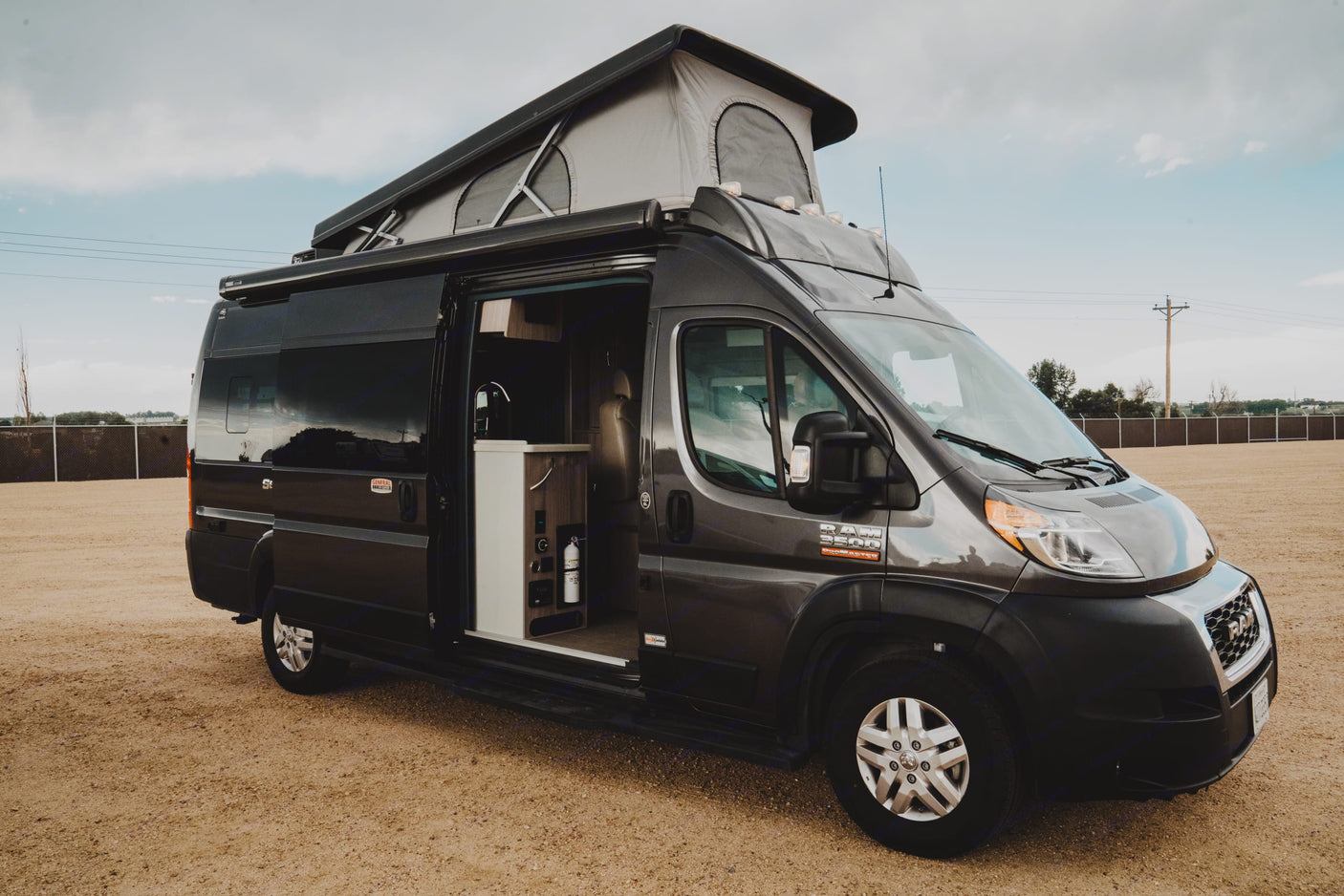 This dark gray pop-top rv rental van, a Ram ProMaster, is ready for adventure with its door open revealing a compact kitchen.