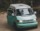 A mint green and white Winnebago camper van **rv rental** on a dirt road with its door open, nestled among trees.