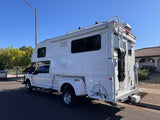 Large white truck camper on a Ford dually with off-road gear, like an RV rental, parked on a sunny residential street.