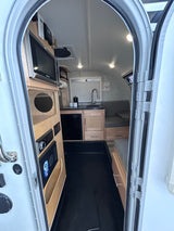 Brightly lit compact RV rental interior, seen from the door. Features a kitchen, dinette, microwave, and fridge.