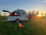 This rv rental adventure features a teardrop trailer, car, and glowing campfire in a grassy field at sunset.