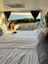 Happy woman enjoying an ocean view from a cozy rv rental bed. Ideal for van life travel & vacations.