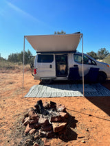 Scenic RV rental: a white camper van with an awning, chairs, and a fire pit set up on red earth under a clear sky.