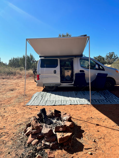 Scenic RV rental: a white camper van with an awning, chairs, and a fire pit set up on red earth under a clear sky.
