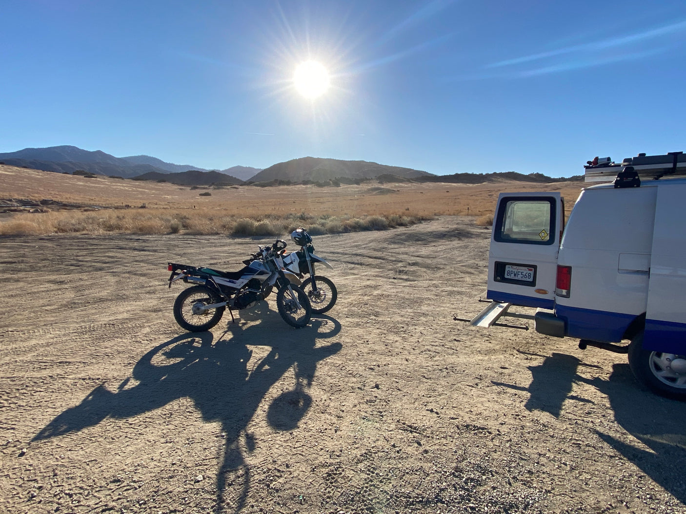 Two dirt bikes await adventure beside an RV rental van with a ramp in a sunny, arid mountain landscape. Ideal for desert exploration.
