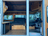 RV rental: Custom camper van interior with warm wood finishes, versatile seating/bed, mini-fridge, and driver's cab.
