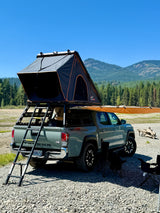 A gray Toyota Tacoma pickup truck with a rooftop tent and awning is set up by a river for camping, an ideal RV rental alternative.