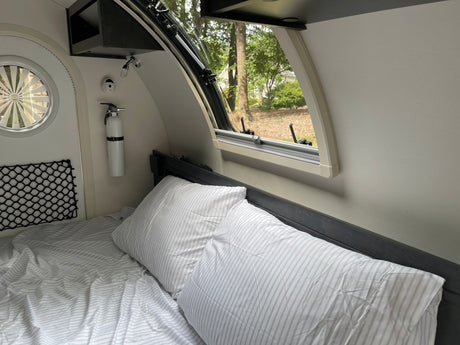 Cozy RV rental interior with a bed, window view of trees, and a fire extinguisher. Ideal for comfortable travel adventures.