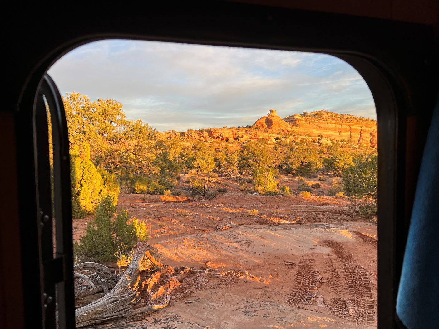 Serene desert landscape viewed from an RV rental. Golden hour light bathes red rocks, desert plants, and tire tracks outside.