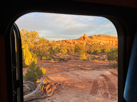 Serene desert landscape viewed from an RV rental. Golden hour light bathes red rocks, desert plants, and tire tracks outside.
