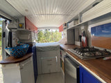 Inside this bright RV rental camper van: A full view of the kitchenette with stove, wooden counter, bed, and decorative blue sink.