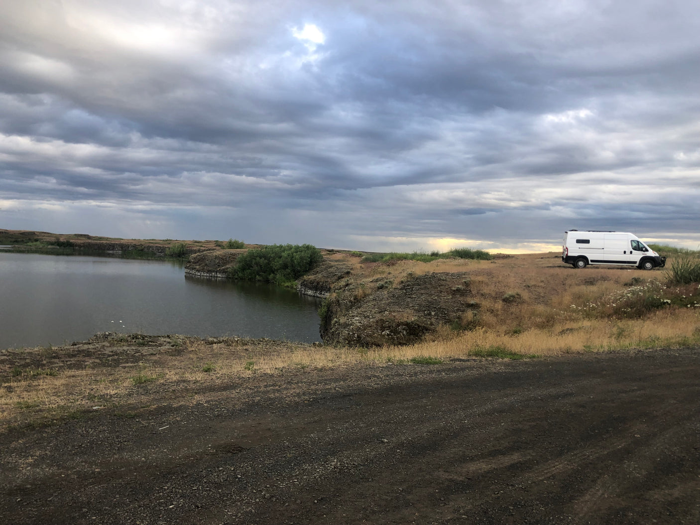 White RV rental parked beside a rugged lake under a dramatic cloudy sky.