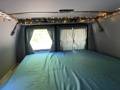 Cozy RV rental interior featuring a bed with blue sheets, charming string lights, and windows with curtains.