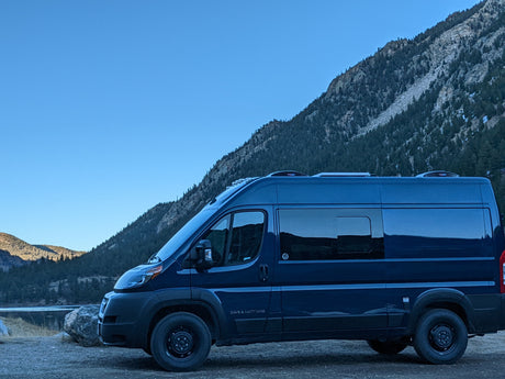 Scenic rv rental: Dark blue camper van parked by a serene lake with a large, tree-covered mountain backdrop.