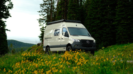 A beige Mercedes-Benz Sprinter RV rental camper van in a mountain meadow with yellow wildflowers and forest.|Explore the Mercedes Winnebago Revel RV rental floorplan. Features a lift bed