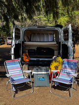 This RV rental camper van set up for adventure: open back reveals a cozy bed, guitar, cooler, and chairs for outdoor enjoyment.
