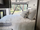 Comfortable RV rental bed with decorative pillows, offering a peaceful forest view through open rear doors. Perfect for glamping.
