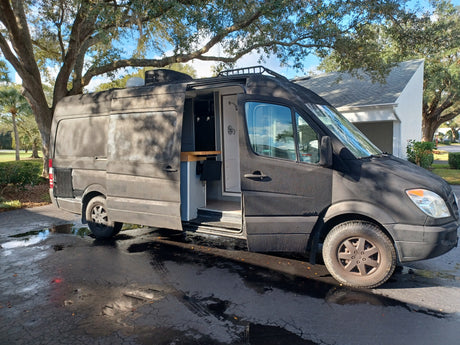 Black custom Mercedes Sprinter camper van, equipped for rv rental. Interior shows wood counter, TV, and an outdoor shower.
