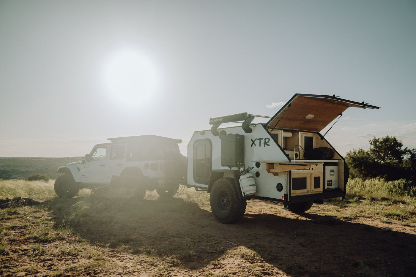 Adventure-ready RV rental: A white Jeep tows an off-road trailer with an open kitchen, bathed in golden light on a dirt path, perfect for overland camping.