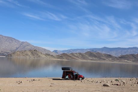 Red camper van, an ideal rv rental, parked lakeside with mountains and blue sky. Awning out for a tranquil outdoor retreat.