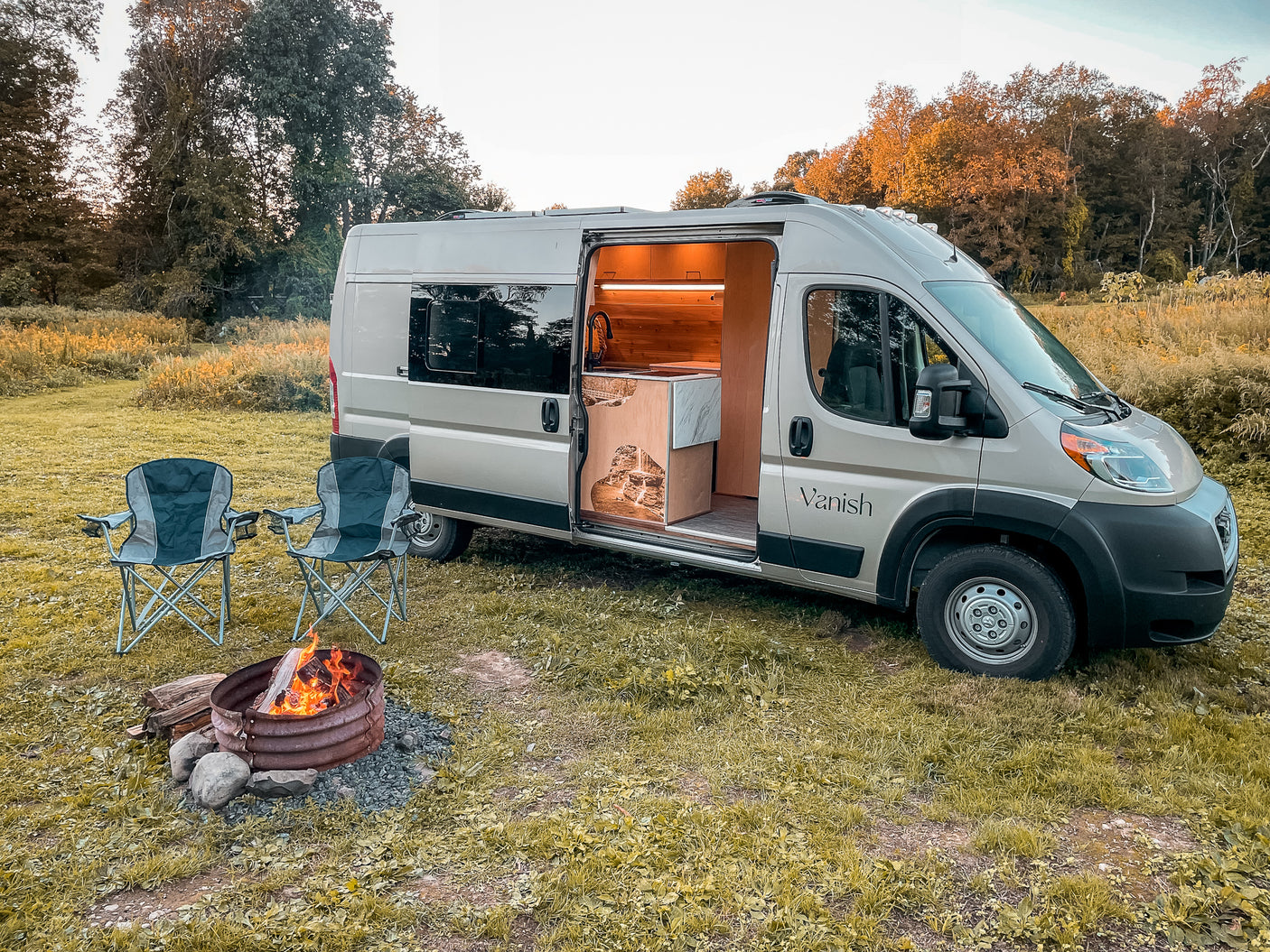 Ready for adventure? This RV rental shows a modern camper van with an open door revealing its kitchen, beside a cozy fire pit & chairs.