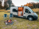 Ready for adventure? This RV rental shows a modern camper van with an open door revealing its kitchen, beside a cozy fire pit & chairs.