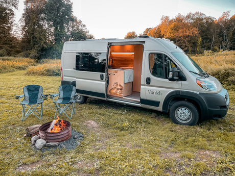 Ready for adventure? This RV rental shows a modern camper van with an open door revealing its kitchen, beside a cozy fire pit & chairs.