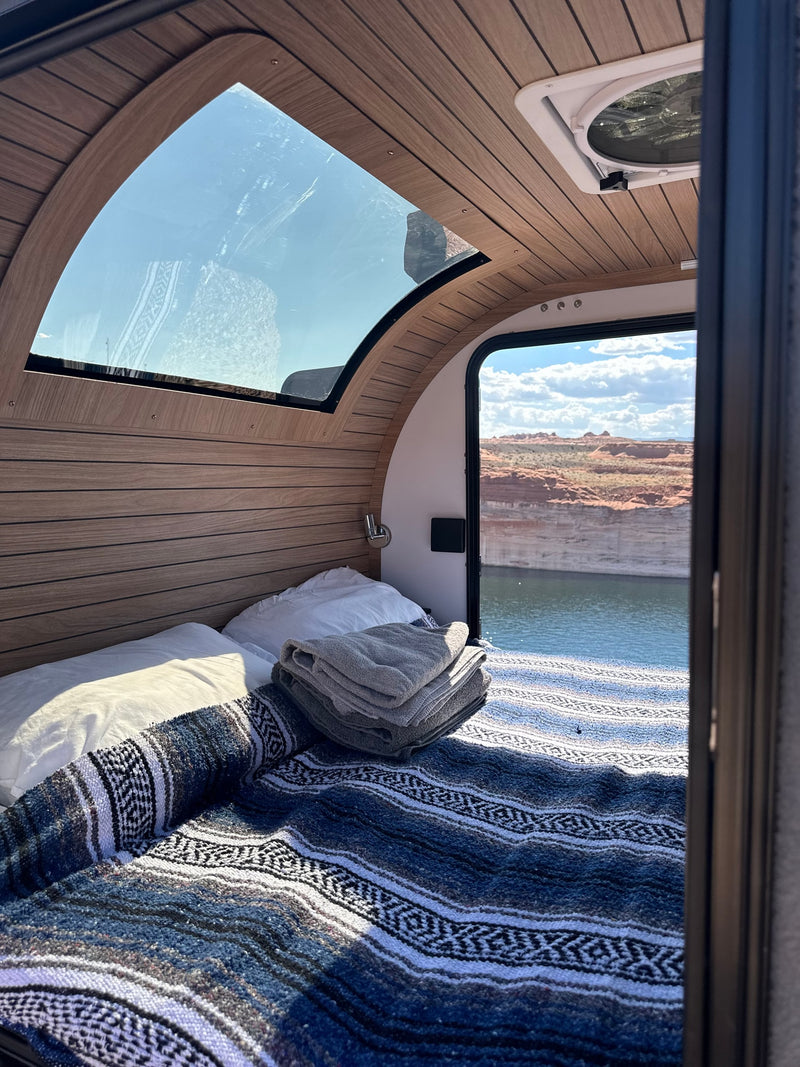 Inside a comfy **RV rental**, a bed with a patterned blanket faces an open door with a beautiful view of a lake and red rock canyons. — photo 6