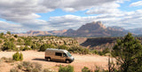 Light-colored RV rental on a dirt road in a vast desert landscape with red rock canyons and majestic mountains.