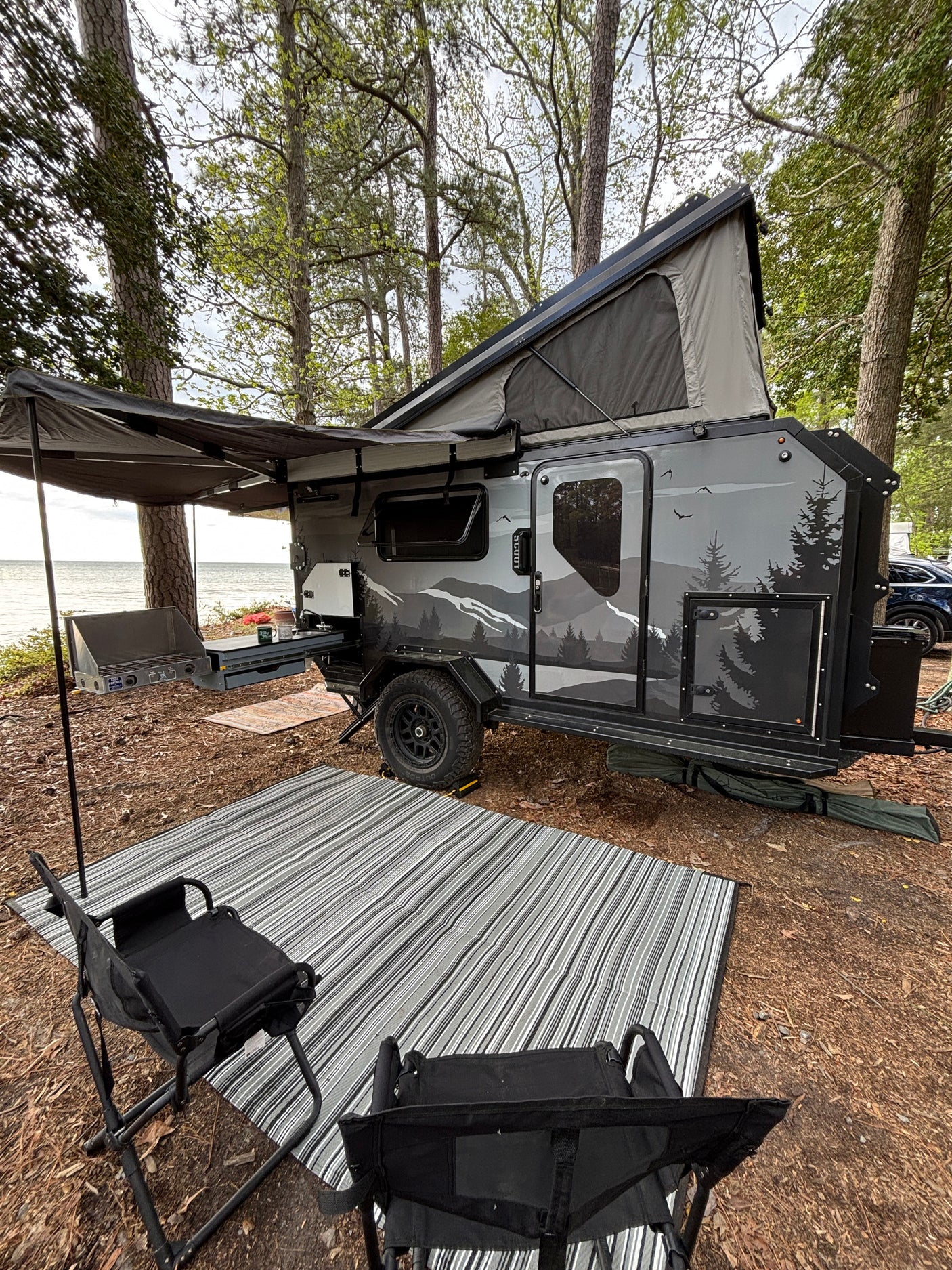 Lakeside RV rental: Modern teardrop camper setup with outdoor kitchen, pop-up tent, awning, chairs, and rug for camping.