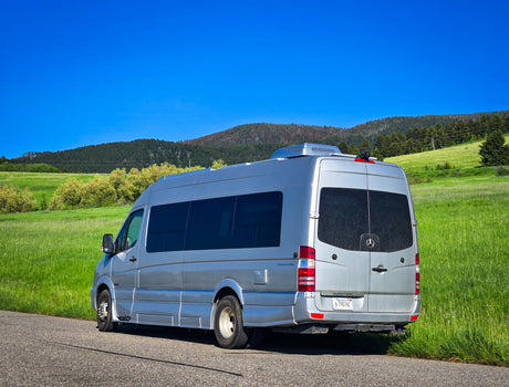 Silver Mercedes Sprinter RV rental van parked on a road with green hills, trees, and blue sky. Ideal for road trips.