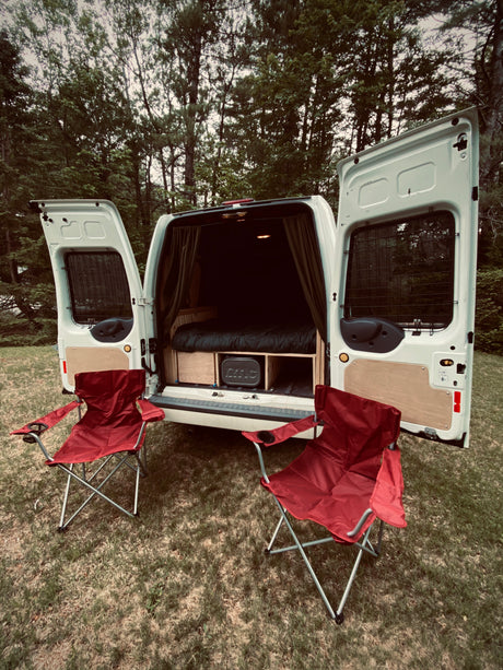 White camper van, customized with bed and storage, opens to two red camping chairs. Perfect for your next RV rental adventure.